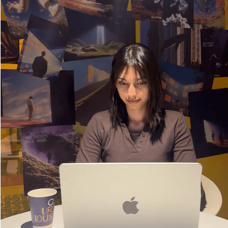 Sanskriti Bhatia (Sans Bhatia) at her desk in Seoul, working on a MacBook in front of a wall of printed images.